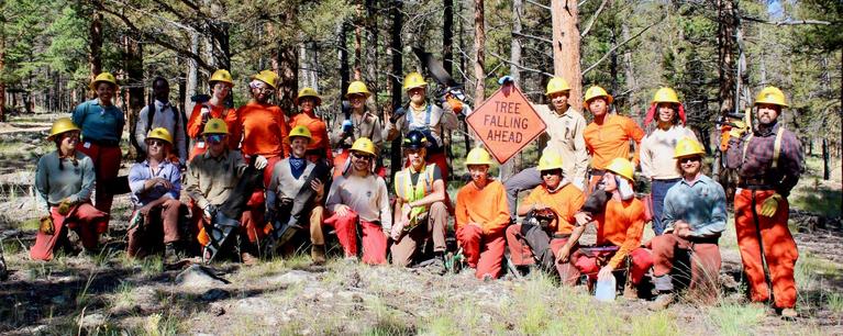 21 corps members from the southwest conservation corps wearing hard hats and chaps, posing in a ponderosa pine forest. 