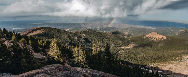 Landscape photo of Pikes Peak in Colorado, with mountains, trees, a cloudy sky, and a rainbow